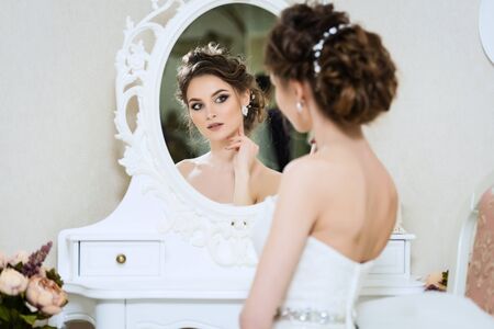 Beautiful young bride in front of the mirror. Portrait with wedding makeup, hairstyle, dressの写真素材