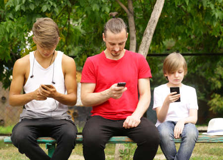 father and two sons sitting on a bench and look at smartphonesの写真素材