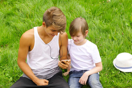 Older and younger brother sitting on the grass and looking to the smartphoneの写真素材