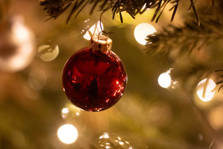 Close-up of red glass Christmas ornament hanging on pine tree with golden lights and festive bokeh