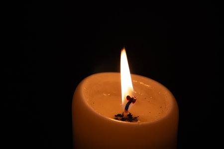 Single burning candle flame glowing in darkness with warm orange wax and black background
