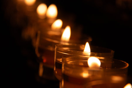 Row of small candle flames glowing in glass holders in a dark setting with soft bokeh effectの写真素材