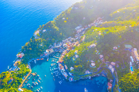 A breathtaking aerial photograph of the beautiful marina in Portofino, Italy, bathed in warm sunlight, showcasing luxury yachts, and the surrounding lush green hillsの写真素材