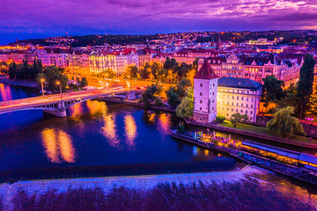 A beautiful view of Prague riverside during a summer evening, showcasing the calm Vltava River, illuminated historical buildings, and vibrant cityscape under the warm night glowの写真素材