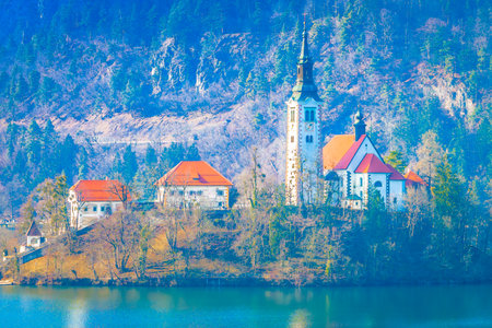Scenic view of the small island in the center of Lake Bled, Slovenia, featuring the iconic church surrounded by crystal-clear alpine waters and dramatic mountainous landscapeの写真素材