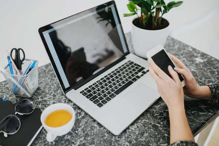 Young woman working from home. Drinking hot tea, holding mobile phone, checking messages.
