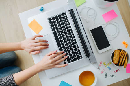 Female workspace desk at home. Woman hands typing on a laptop. Flatlay with mobile phone, headphones, white mug, stickers ans cookies. Hipster working from home.の写真素材
