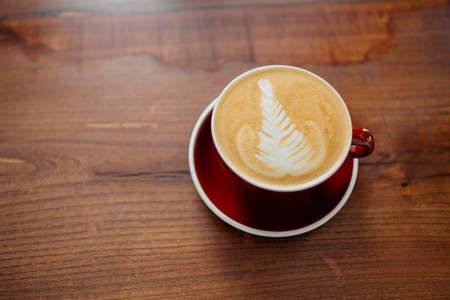 A cup of cappucino in a red cup on a sunny morning, waiting on a wooden table to be tasted.