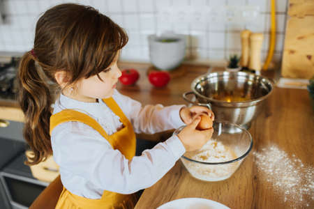 A little girl helping mom to bake a cake in the kitchen. Smashing eggs into a bowl with flour.の写真素材