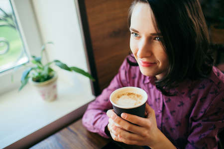 Close up overview of an attractive young woman drinking delicious cappuccino in a coffee shop, smiling.の写真素材