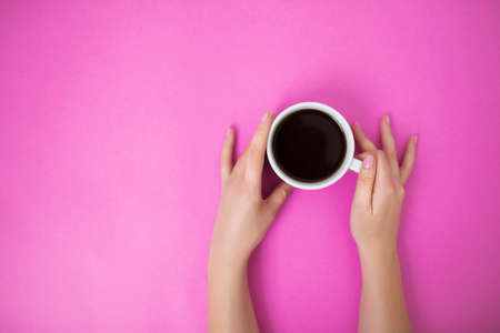 Flatlay with black coffee in a white mug on a pink background. Woman hands with manicure gently holding cup. Copy space.の写真素材