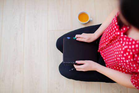 Overview of a woman sitting on the floor, getting ready to write. Female hands holding a black notepad, a cup of tea beside.の写真素材