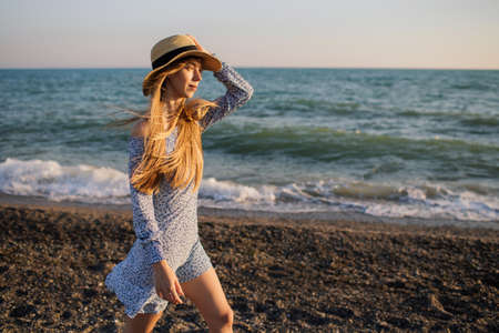 Beautiful young girl in a hat and light blue dress walking along the beach at sunset.の写真素材
