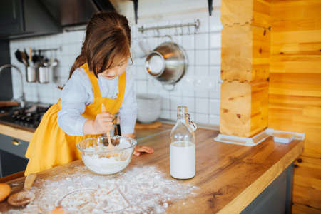 A little cute girl getting ready to bake a cake in the kitchen. Stirring dough in a bowl. Flour and milk on the table.の写真素材