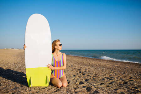 Young fit surfer girl sitting on the sand at the beach with a surf board. Looking towards the water.の写真素材