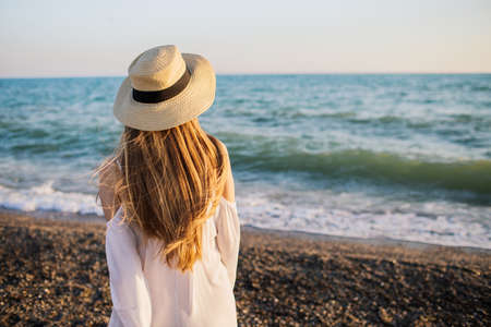 Young blonde girl in a straw hat looking at the sea waves. Back view.の写真素材