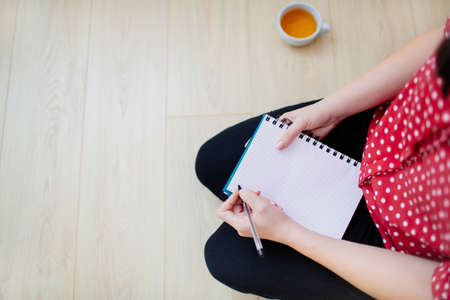 Top view of a woman sitting on the floor, getting ready to write in a notepad. Female hands holding a pen and notepad, a cup of tea beside.の写真素材