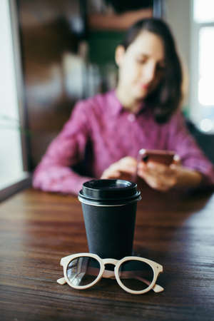 Young woman scrolls her smartphone, sitting in a cafe. A cup of coffee and sunglasses beside.の写真素材