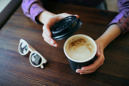Female hands holding a cup of cappuccino in black takeaway paper cup. Coffee take away.の写真素材