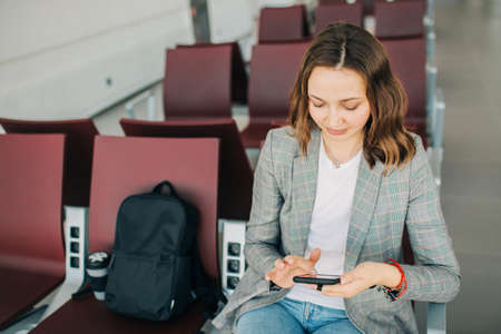 Young attractive girl at the airport waiting for her departure. Scrolling smartphone. Coffee cup and backpack beside.の写真素材
