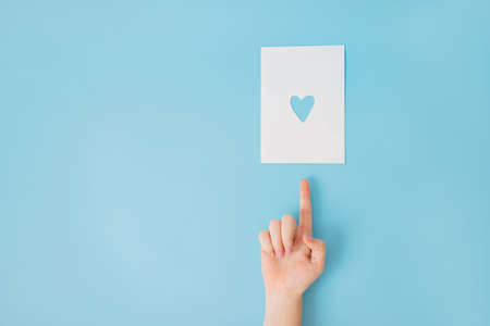 Female hand with manicure on a blue background pointing at a white sheet of paper with heart shaped hole in it.の写真素材