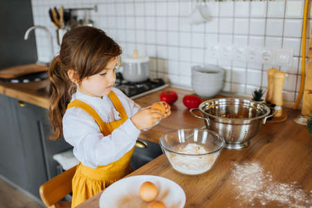 Little cute girl helping mother to bake a cake in the kitchen. Smashing eggs into a bowl with flour.の写真素材