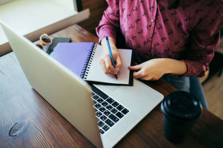 Overview of a young businesswoman writing something down into the notepad, working in cafe. A cup of...