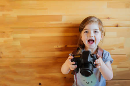 Happy excited little girl with photocamera on light wooden background.