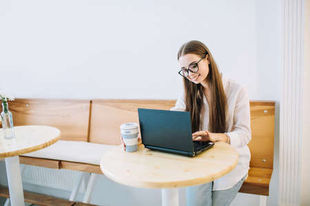 Young smiling woman working on laptop at a cafe, while drinking coffee. Young attractive girl sitting in a coffee shop with black laptop.の写真素材