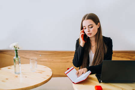 Young successful businesswoman talking on the phone sitting in front of the laptop in a cafe. Preparing to write down information in a notepad.の写真素材