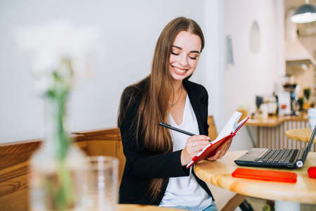 Young smiling businesswoman making notes in her notepad, sitting in front of the laptop in a cafe. Preparing to write down information.の写真素材