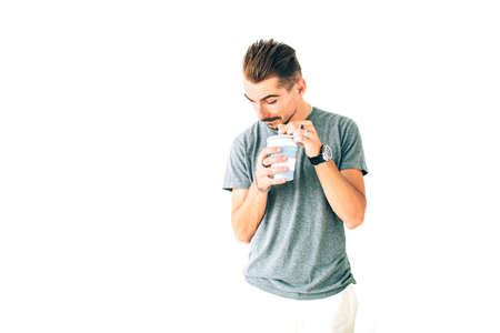 Happy young playful guy in grey t-shirt holding a cup of coffee on white isolated background, peeking inside the cup smiling.の写真素材