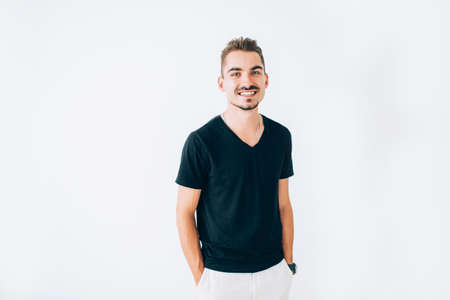 Happy young guy in black t-shirt posing on white isolated background. Standing with hands in the pockets, smiling.の写真素材