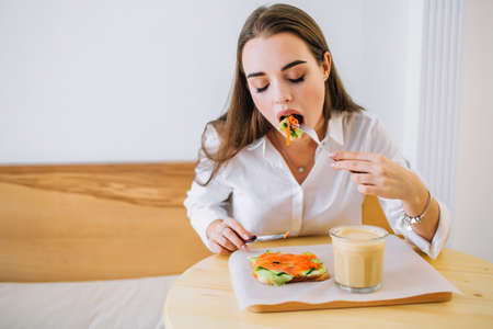 Young attractive woman eating sandwich with salmon and cucumber, sitting at the table with coffee...