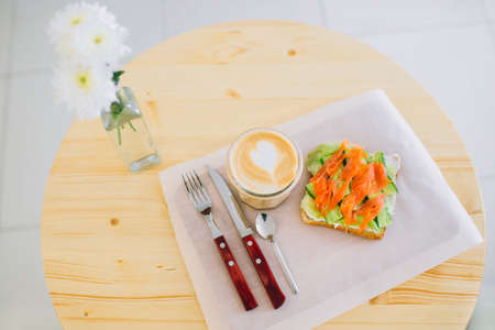 Top view of cappuccino and sandwich with salmon and cucumber waiting on the table. Spoon, fork and a knife beside.の写真素材