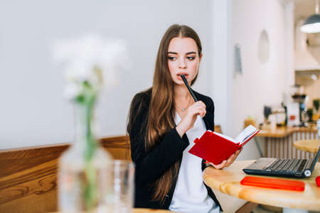 Young concentrated businesswoman holding her notepad and pen, sitting in front of the laptop in a cafe.の写真素材