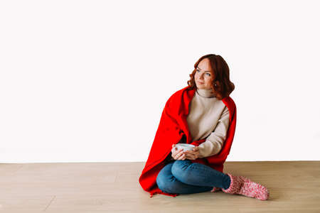Dreaming young red haired woman sitting on the floor isolated on white background. Female model smiling looking up, holding a mug with coffee.の写真素材