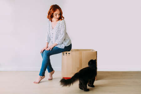 Young beautiful girl moving in the new apartment. Sitting on a carton box on a white background,...