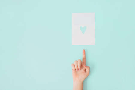 Female hand with manicure on a bleached coral background pointing at a white sheet of paper with heart shaped hole in it.の写真素材