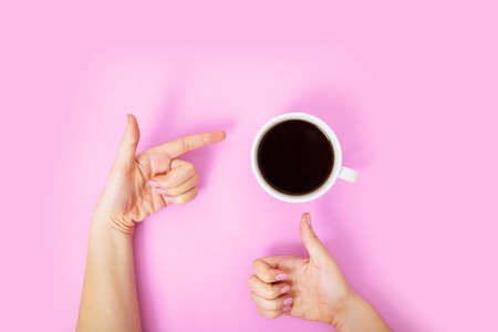 Flatlay with black coffee in a white mug on a pink background. Woman hand showing thumb up sign, and pointing finger at cup with another hand.の写真素材