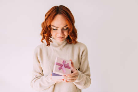 Young girl with festive mood gently smiles, holding a pastel coloured pink gift box with a ribbon. Happy woman, peeking inside a gift box.の写真素材