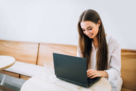 Young smiling girl working on laptop in a cafe, typing on a keyboard. Looking into the screen, pleased and joyful.の写真素材