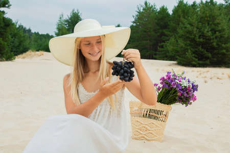 Beautiful girl in a summer hat in nature with a bouquet of flowers eats grapesの写真素材