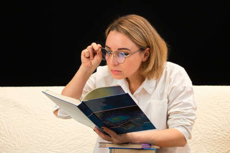 A woman in glasses and a white shirt sits on a sofa, carefully looks into a book.の写真素材