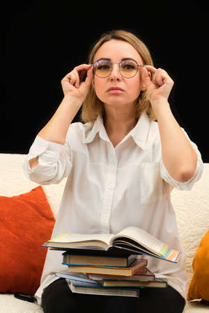 The teacher in glasses and a white shirt sits on the couch, holds books, looks ahead attentively