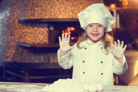 A little girl, soiled in flour, dressed in a chefs uniform and wearing a big white cap, spoils herself by stretching her arms forward, showing her soiled hands.の写真素材
