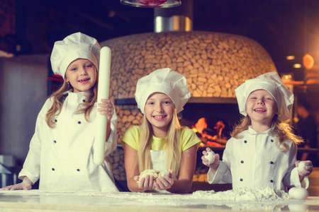 Near the oven, three cheerful little girls, soiled in flour and dressed in chefs uniforms, are preparing a flour product.の写真素材