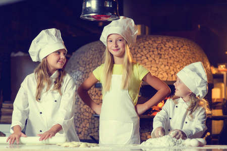 Near the oven, three cheerful little girls, soiled in flour and dressed in chefs uniforms, are making pizza.の写真素材