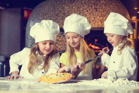 Near the oven, three cheerful little girls, soiled in flour and dressed in chefs uniforms, are making pizza.の写真素材