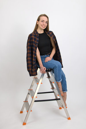Beautiful blue-eyed smiling girl with long blond hair and bare feet expresses emotions of satisfaction while posing while sitting on a folding aluminum ladder. Close-up on a white background.の写真素材
